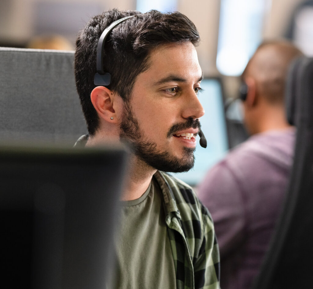 Hispanic man using a computer and talking to a customer over a headset while doing customer support in a call centre.